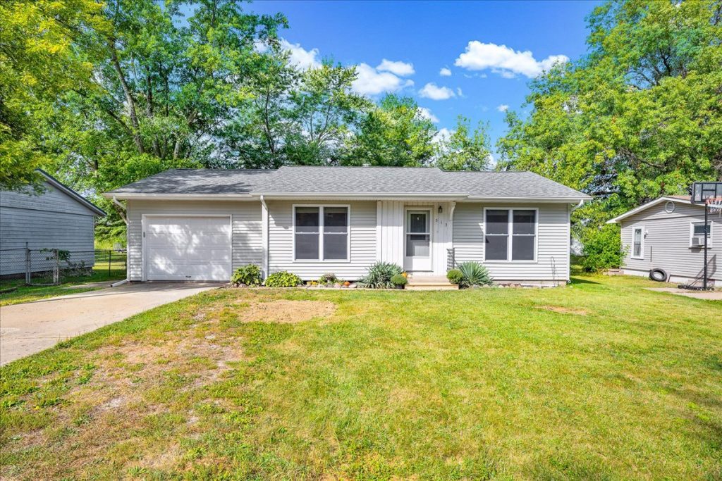 The front of a Ranch style home with attached 1 car garage, white siding, large yard.